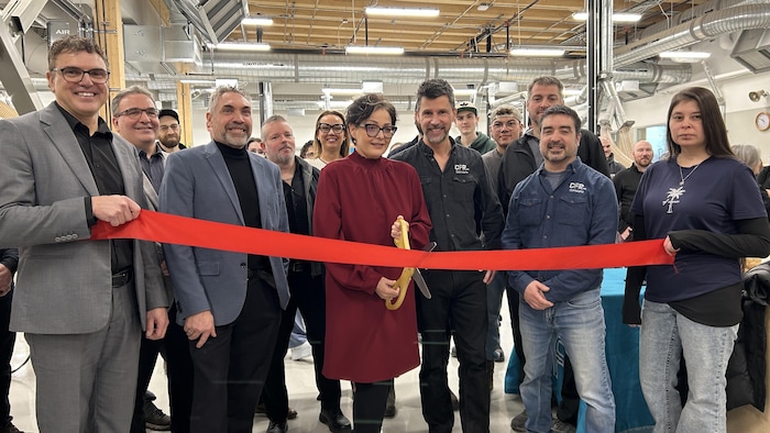 Un groupe de personnes est réuni dans un atelier industriel pour une coupure de ruban rouge. Au centre, une femme tient de grands ciseaux dorés, entourée d'hommes en tenue civile et d'employés portant le logo « CFP Alma ».
