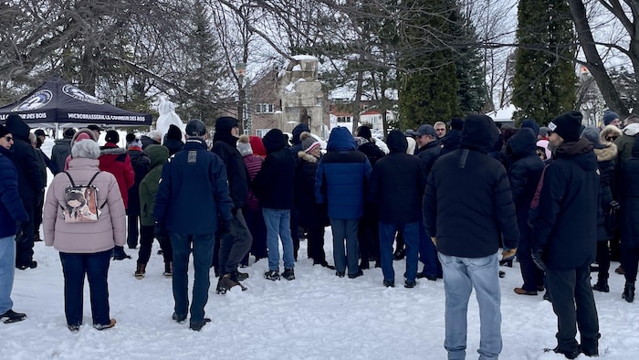 Un grand groupe de personnes, la plupart tournant le dos à la caméra, est rassemblé sur un sol enneigé dans un parc. Les participants portent des vêtements d'hiver. Une tente noire est visible à gauche.