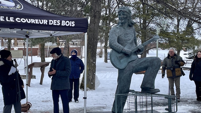 Une statue en bronze d'un homme jouant de la guitare, juché sur un chariot d'épicerie, au centre d'un parc enneigé. À gauche, un petit groupe de personnes est rassemblé sous une tente noire.