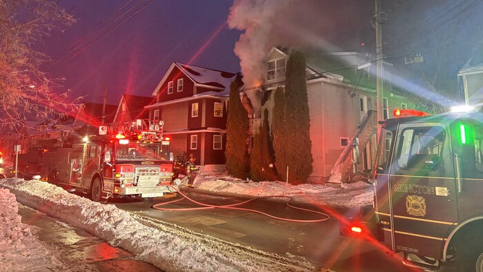 De la fumée noire et des flammes sortent d'une fenêtre d'un duplex. Des camions de pompiers sont garés devant le logement.