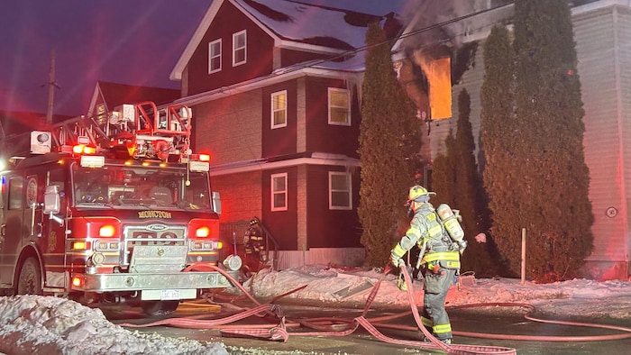 Une maison en proie aux flammes en arrière-plan. Au premier plan, un pompier s'active à côté d'un camion.