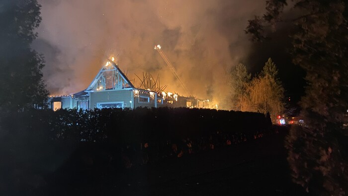 Dans la nuit, la grande échelle des pompiers est déployée au-dessus d'un garage en flammes dont le toit s'est effondré. 