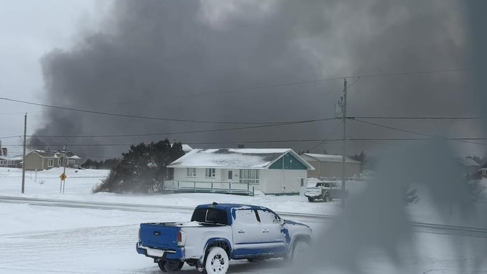 Un panache de fumée s'étend dans le ciel de Gros-Cap, derrière une route et des habitations enneigées.