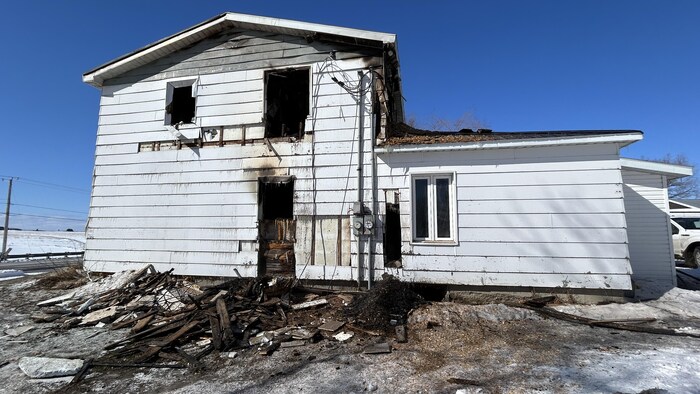 Cette photo montre l'extérieur d'une maison blanche à deux étages lourdement endommagée par le feu. Les fenêtres sont calcinées, une partie du revêtement est arrachée et un amas de débris brûlés jonche le sol enneigé devant la structure.