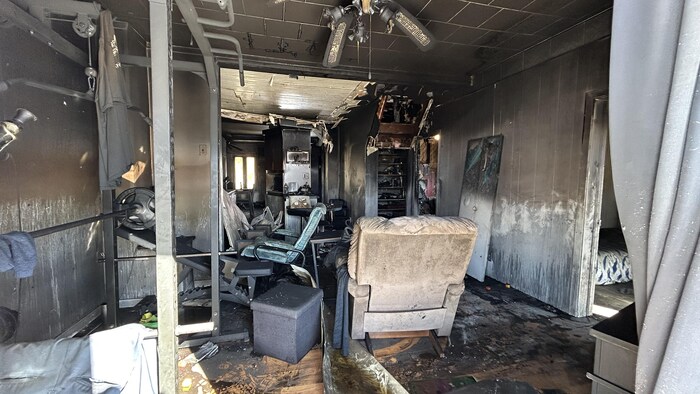 Cette photo montre l'intérieur d'une habitation gravement endommagée par un incendie. Les murs et le plafond sont recouverts de suie noire. On aperçoit un ventilateur de plafond fondu, un appareil de musculation, un fauteuil et des débris épars.
