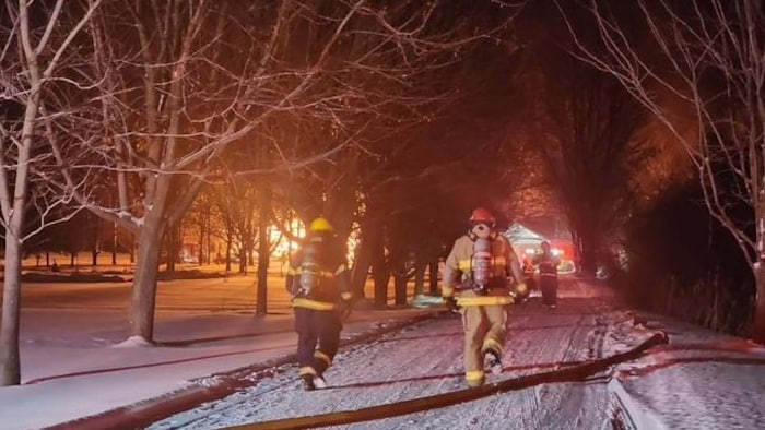 Des pompiers marchent dans une entrée enneigée bordée par des arbres, en direction d’une maison en flammes.
