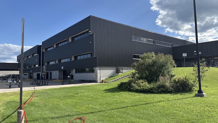 Un grand bâtiment moderne au revêtement sombre s'élève derrière une pelouse verdoyante. À droite, un mât arbore le drapeau du Québec. Le premier plan est marqué par un poteau entouré d'un ruban de sécurité rouge sous un ciel nuageux.