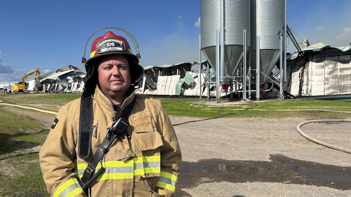 Un pompier en uniforme et casque rouge pose au premier plan face à l'objectif. Derrière lui se trouvent deux grands silos métalliques et les restes effondrés d'un bâtiment agricole sous un ciel bleu dégagé.