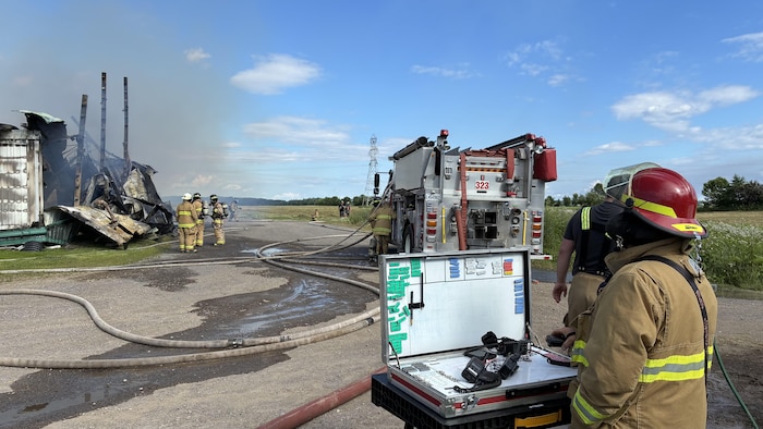 Des pompiers interviennent sur les décombres fumants d'un bâtiment détruit. Un camion de pompiers est stationné à côté de longs tuyaux d'arrosage au sol. Au premier plan, un pompier observe la scène près d'une mallette de commandement ouverte.