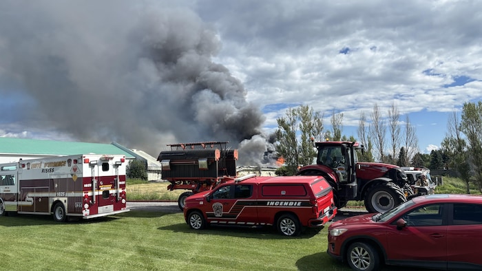 Une épaisse colonne de fumée noire s'élève d'un bâtiment en feu en arrière-plan. Au premier plan, plusieurs véhicules d'urgence rouges, dont une unité de soutien et un pick-up "INCENDIE", sont stationnés sur l'herbe aux côtés d'un tracteur agricole.