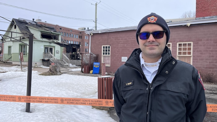 Un homme en uniforme de pompier sourit à la caméra. Un ruban de sécurité de la Sûreté du Québec et une maison calcinée sont visibles derrière.