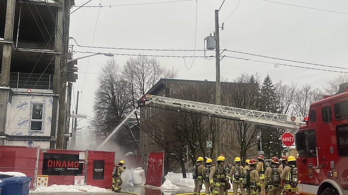 Des pompiers debout devant l'immeuble. Une lance d'incendie projette de l'eau.