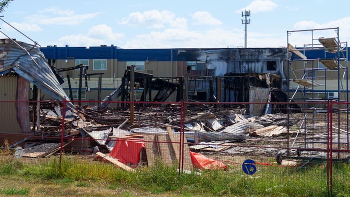 Des restes calcinés d’un bâtiment détruit par un incendie, avec des débris éparpillés derrière une clôture de chantier.