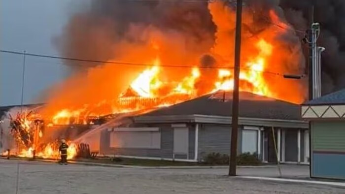 Des flammes dépassent du toit du bâtiment accompagnées par de la fumée noire. Un pompier avec une lance à eau tente de venir à bout du brasier. 