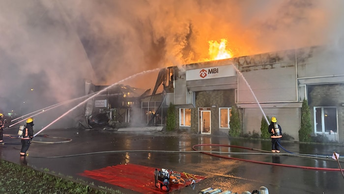 Des pompiers essaient d'éteindre un incendie. On voit les flammes s'élever du toit d'un bâtiment.