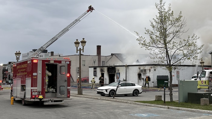 Un pompier en haut d'une échelle sur un camion éteint un brasier avec un puissant jet d'eau. Des pompiers sont à côté du bâtiment qui dégage de la fumée.