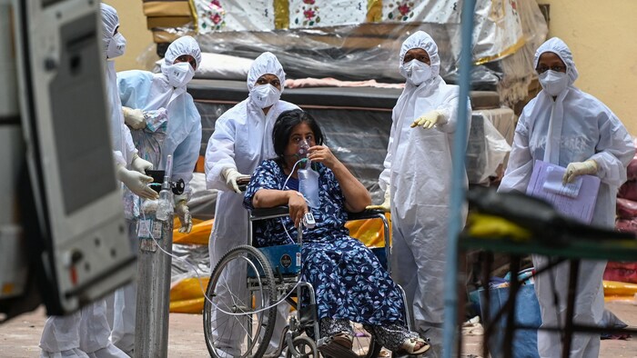 La femme est assise dans une chaise roulante et utilise un masque à oxygène. Elle est entourée de plusieurs personnes portant des habits de protection.