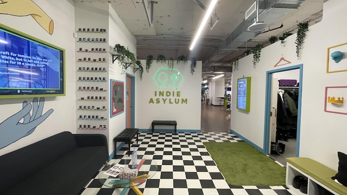 L'entrée de l'Indie Asylum, avec un plancher quadrillé blanc et noir, des écrans, un tapis en gazon synthétique, et une manette de jeux vidéo en néon au mur.