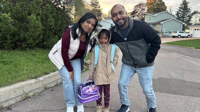 La fillette porte un sac à dos et tient une boîte à diner.  Elle est entourée de sa mère Khusboo et de Vimal.