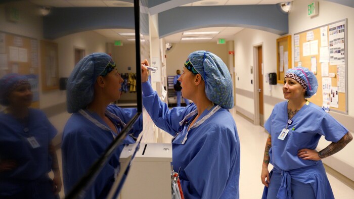 Une infirmière de profil écrit sur un tableau pendant que sa collègue la regarde.