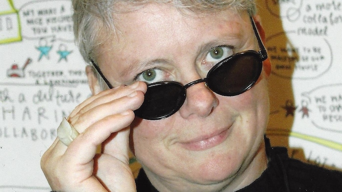 Une femme sourit en baissant ses lunettes fumées lors d'une conférence.