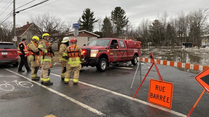 Cinq pompiers sont devant une voiture du service de sécurité incendie, stationnée près d'une rue inondée. 