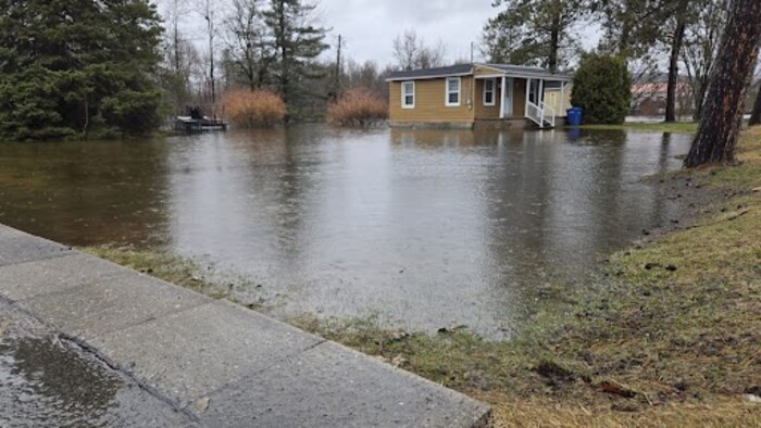De l'eau a débordé sur un terrain, encerclant une petite maison. À l'avant-plan, un trottoir est dégagé. 