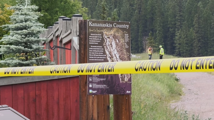 Un ruban de signalisation jaune interdisant l'accès est tendu devant un panneau d'information de Kananaskis Country, alors que deux personnes portant des gilets de haute visibilité se tiennent sur un chemin en arrière-plan.