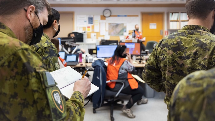 Une salle de réunion avec des gens assis devant des ordinateurs sont en compagnie de soldats des Forces armées canadiennes.