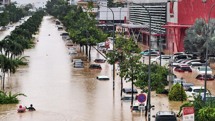 Deux personnes marchent dans des rues inondées. L'eau monte jusqu'à leur taille, et plusieurs véhicules sont immobilisés le long de la rue, englouties presque en totalité dans l'eau.