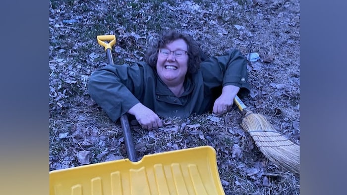 Christine Keilback, amusée, se cramponnant à un balai et une pelle, le corps dans un trou.