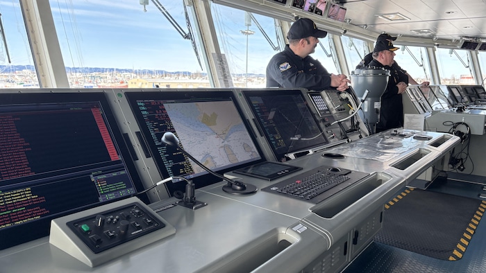 Des officiers de la marine sont à l'intérieur d'un navire, où on voit des consoles de navigation.