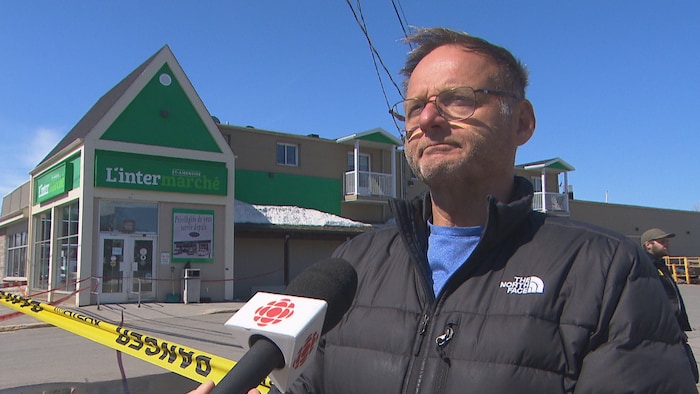 Un homme portant une veste The North Face est interviewé par Radio-Canada devant un magasin L'Intermarché à Saint-Ambroise. Le périmètre est délimité par du ruban jaune « DANGER ». On aperçoit de la neige accumulée sur le toit du bâtiment.