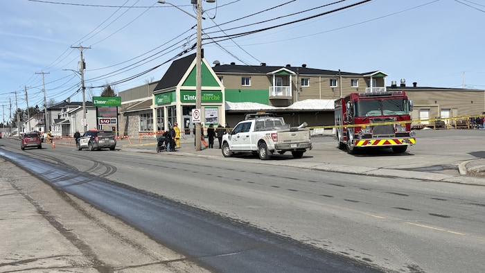 Une scène d'urgence se déroule devant une épicerie L'Intermarché. Un camion de pompiers, une autopatrouille et un véhicule technique sont stationnés près d'un cordon de sécurité jaune. Plusieurs personnes et intervenants observent la situation sous un ciel dégagé.
