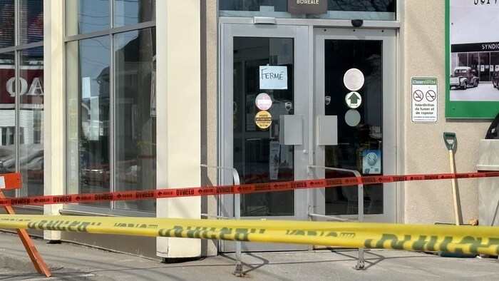 Cette photo montre l'entrée d'un commerce portant une affiche manuscrite « FERMÉ ». Des rubans de sécurité jaunes et rouges barrent l'accès au bâtiment. On aperçoit également un logo « Zone Boréale » au-dessus des doubles portes vitrées.