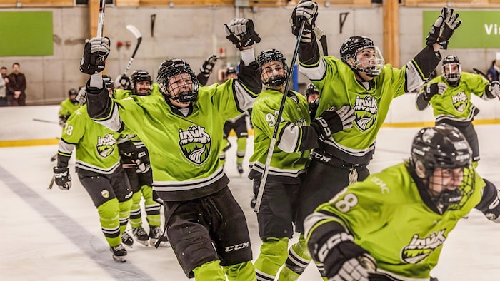 Plusieurs joueurs de hockey en uniforme vert lime et noir célèbrent sur la glace, les bras levés et les bâtons hauts. Leur chandail arbore le logo « Inuk ». En arrière-plan, on aperçoit une affiche « UQAC » dans un aréna au décor industriel.