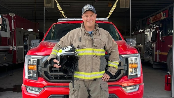 Un pompier se tient debout avec son uniforme et il tient un casque d'une main, en souriant.