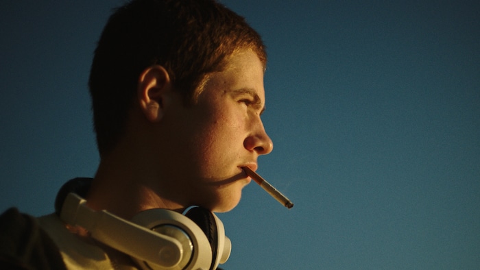 Un jeune garçon fume une cigarette à l'extérieur, devant un ciel complètement bleu. 