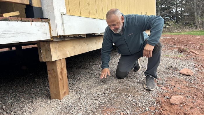 Un homme pose un genou au sol et regarde en dessous de la fondation de son chalet, posé sur des grosses poutres en bois.