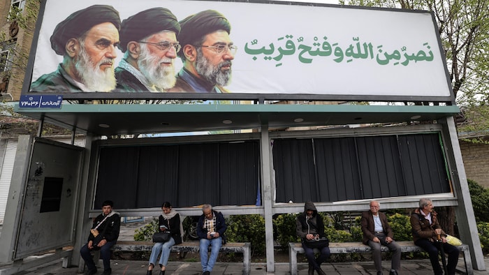 Des Iraniens sont assis à un arrêt de bus sous un panneau d'affichage représentant le défunt guide suprême iranien, l'ayatollah Rouhollah Khomeini (à gauche), son successeur, le défunt ayatollah Ali Khamenei (au centre), et le nouveau guide suprême Mojtaba Khamenei, à Téhéran, le 31 mars 2026.