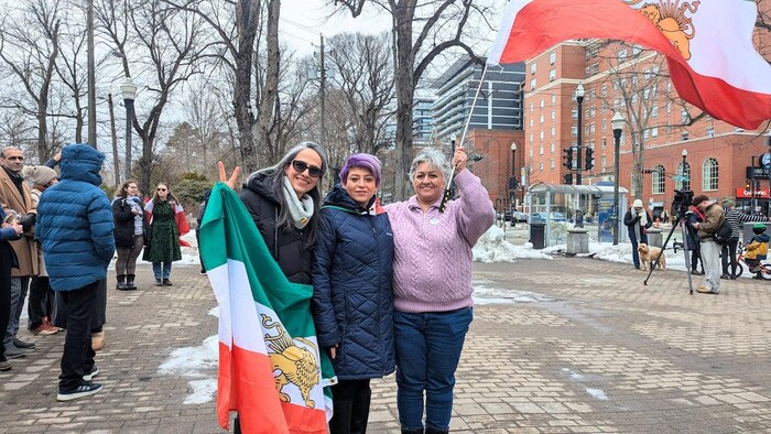 Trois femmes posent ensemble sur une place publique. Deux d'entre elles agitent des drapeaux de l'Iran.