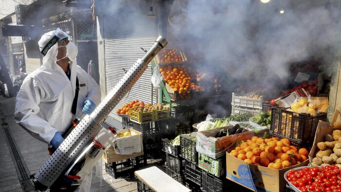 Un homme portant un équipement de protection et un masque vaporise du désinfectant au-dessus d'un étalage de fruits et légumes.