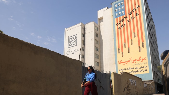 Une femme iranienne en chemise bleue marche à côté d'une bannière de propagande anti-américaine.