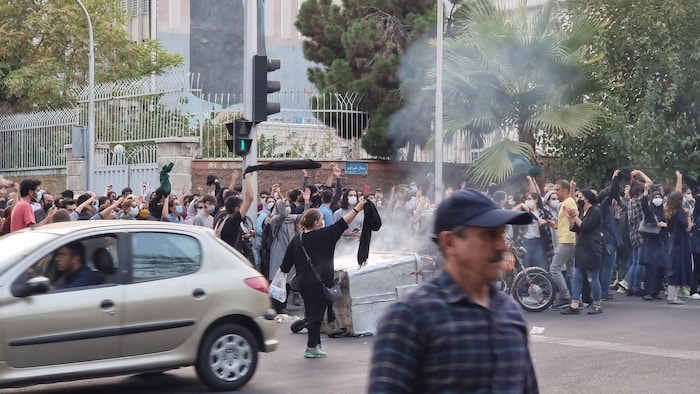 Des manifestants, jeunes pour la plupart, et comprenant des femmes n'arborant pas le hijab, se massent dans la rue, dans un nuage de fumée ou de poussière.