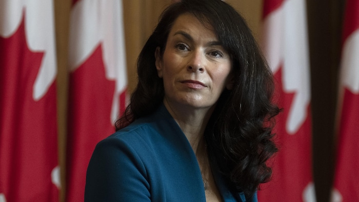 Une femme aux cheveux longs et noirs regarde de côté avec des drapeaux du Canada à l'arrière-plan.