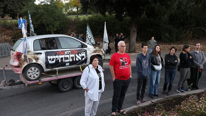 Des Israéliens se tiennent près d'une voiture représentant symboliquement l'attaque du Hamas du 7 octobre 2023, à l'occasion de son troisième anniversaire, devant la Knesset, le parlement israélien à Jérusalem, le 7 octobre 2026.