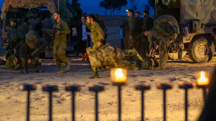 Des chandelles à l'avant-plan de l'image et à l'arrière-plan, des soldats israéliens vêtus de leur treillis, et des camions militaires de couleur kaki. 
