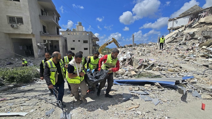 Des secouristes transportent une dépouille dans les décombres, tandis qu'une pelle mécanique dégage les débris après une frappe israélienne.