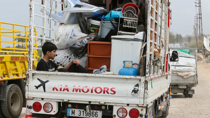 Un adolescent assis à l'arrière d'une camionnette de marque Kia, dans laquelle sont entassés des meubles et des affaires.