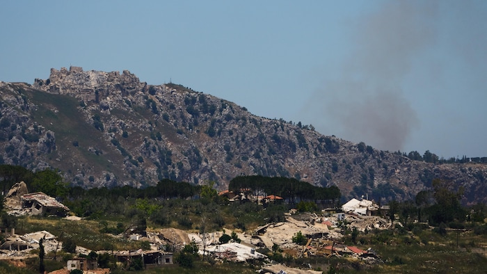 Un paysage montrant un éperon rocheux sur lequel se trouvent les ruines d'un château, des habitations détruites et de la fumée qui s'élève à droite de l'image.